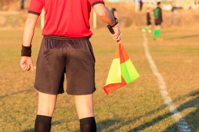 Image of the back of a linesman standing at sidelines of football pitch with players in the distance. 