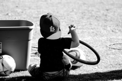 Black and white image from behind of boy sitting on grass pitch next to footballs. 