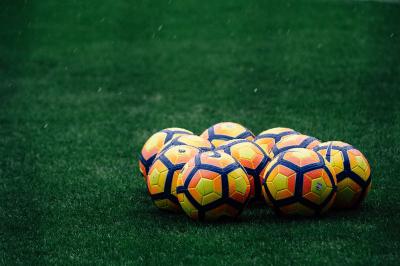 Image of nine footballs grouped together on the ground of a grass pitch.