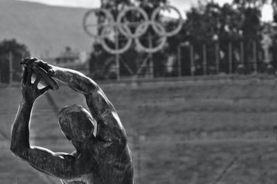 Black and white image of the back of a statue of an athlete, with sculpture of the olympic rings in the distance.